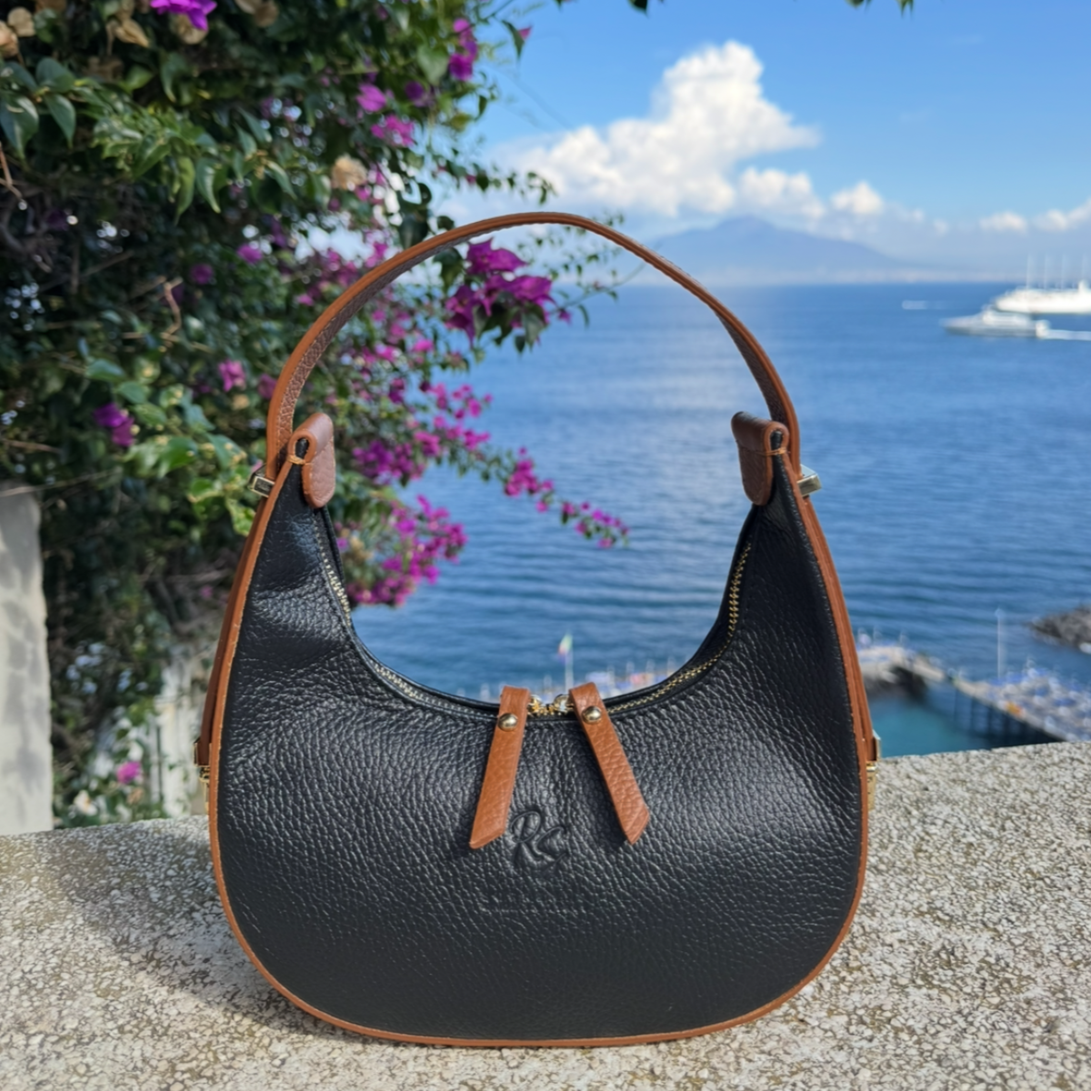 Black leather handbag with brown accents on a ledge with a scenic background of water and flowers.