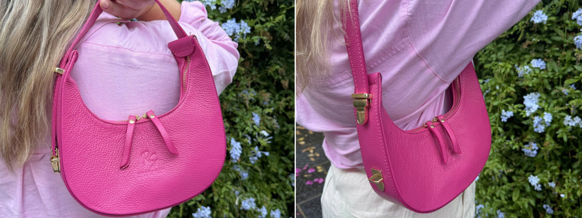 A bright pink pebbled leather crescent shoulder bag with gold-tone hardware, shown worn crossbody by a model in a pink top against a background of green foliage and small blue flowers.